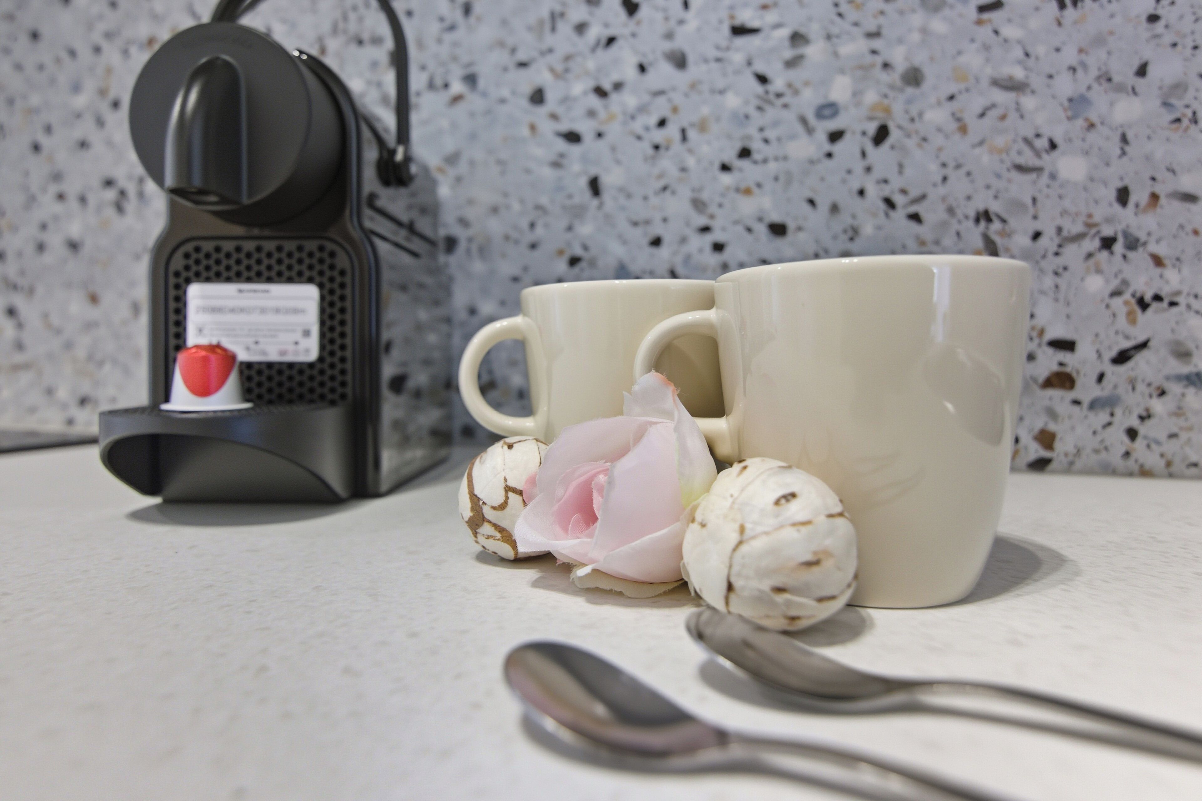 A kitchen counter with a coffee maker, two mugs, a flower, and a spoon.