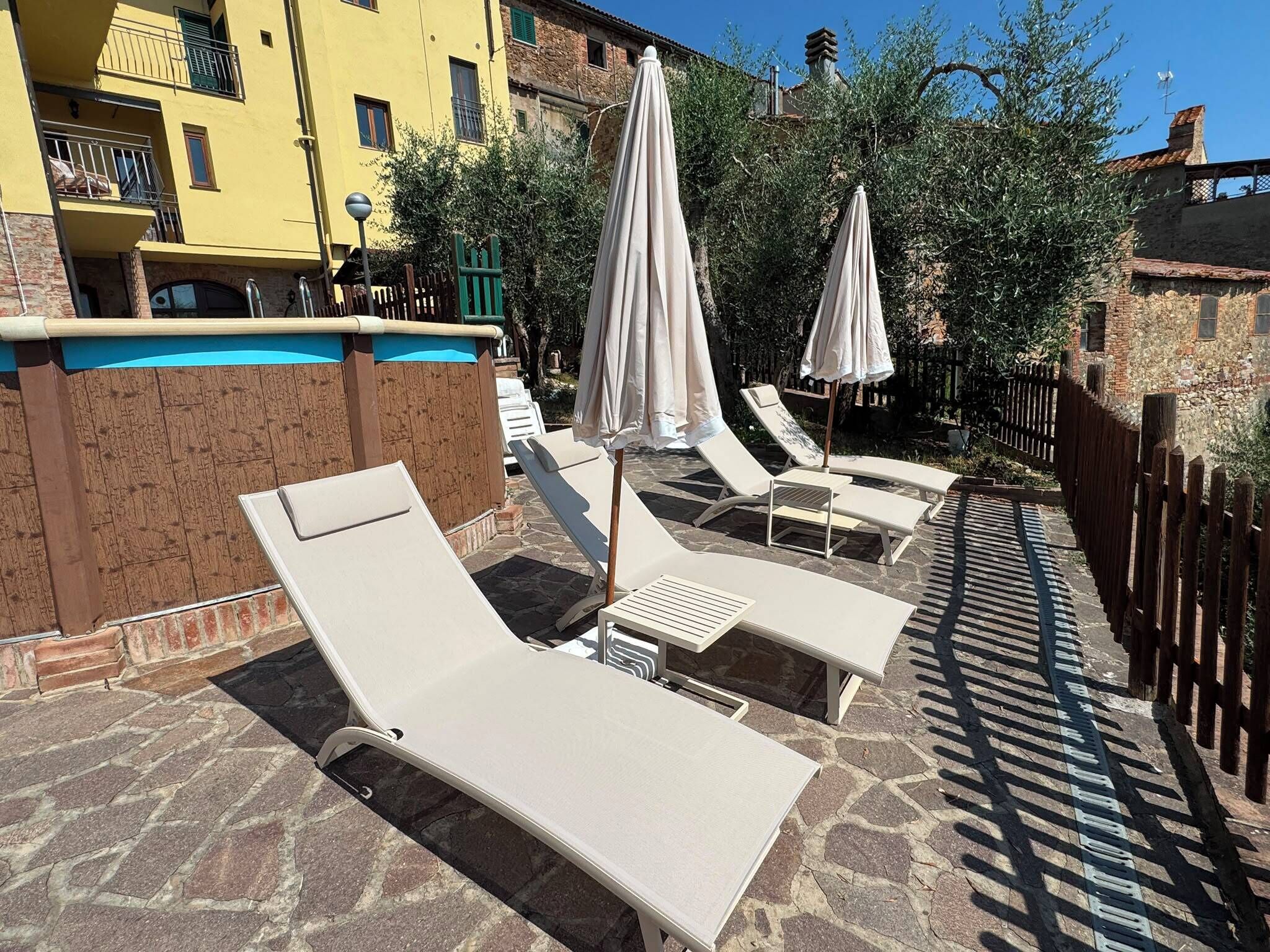 A poolside area with lounge chairs and umbrellas, stone flooring, and a wooden fence.