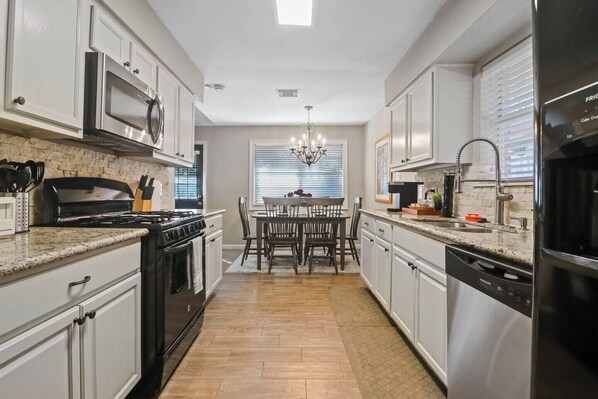 A modern kitchen with stainless steel appliances, a dining area with a chandelier, and a view through the window. - Woodlands 1 Story Retreat • 3BR • Game Room • Gym • Fenced Yard • Dining for 6 (Spring)
