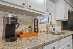 Une cuisine équipée d’une machine à café Keurig, d’un évier et d’une fenêtre avec des stores.
