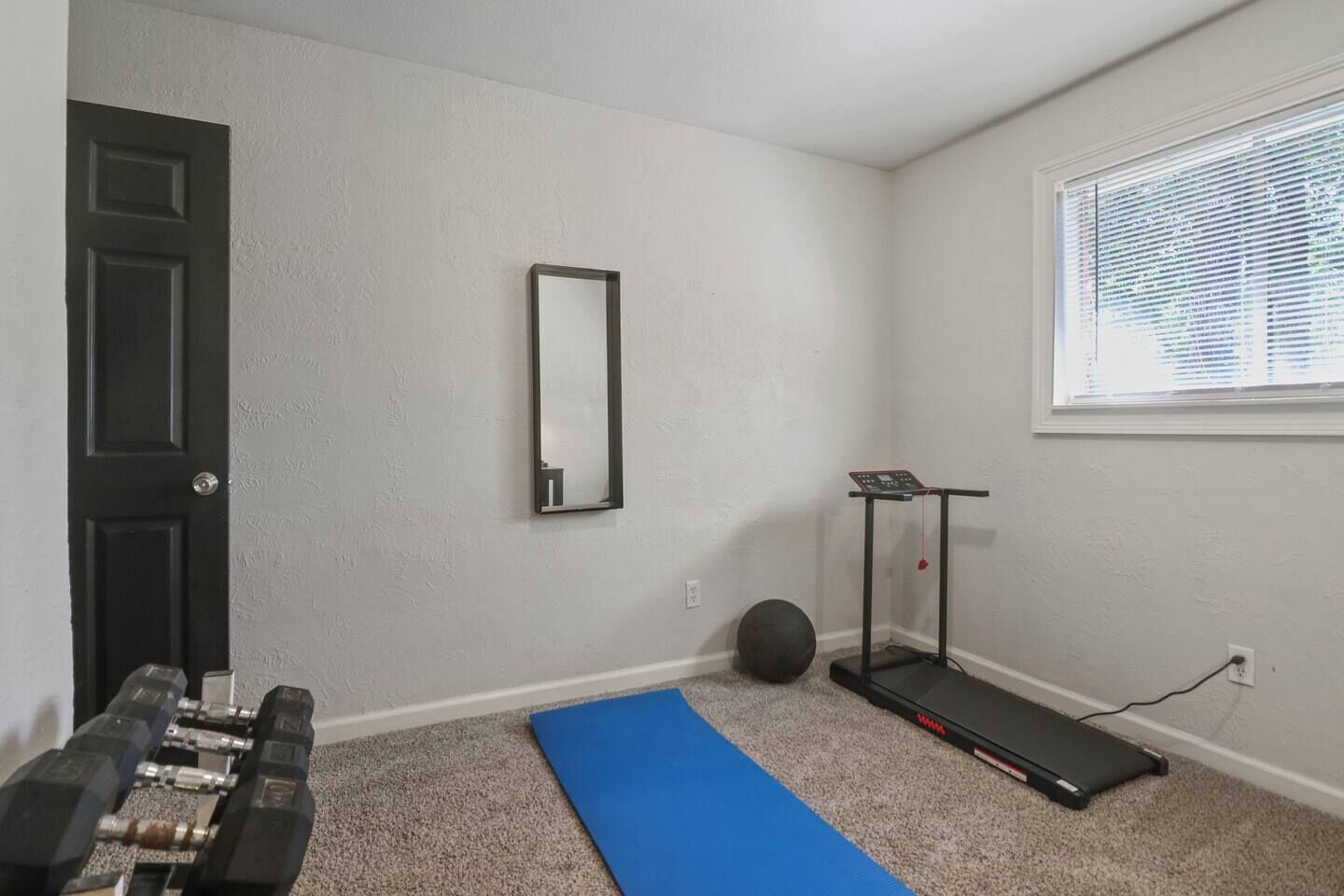 A room with a door, a mirror, a treadmill, dumbbells, a blue mat, and a window with blinds.