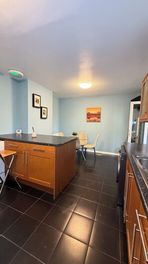 A kitchen with dark tiled flooring, wooden cabinets, a black countertop, and a recessed ceiling light. - Cosy 5-bed house in Swanley (Swanley)