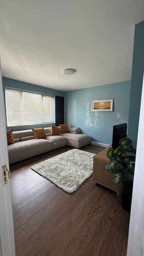 Uma sala de estar com sofá modular, tapete branco, planta e um quadro na parede.