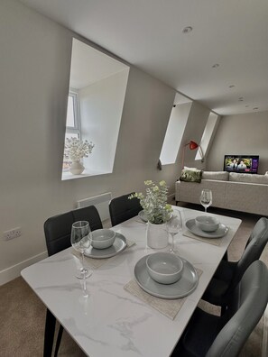A modern dining area with a white table set for four, a large mirror on the wall, and a television in the background.
