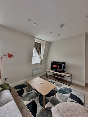 A modern living room with a television displaying a Netflix interface, a red floor lamp, a small wooden table, and a patterned rug.