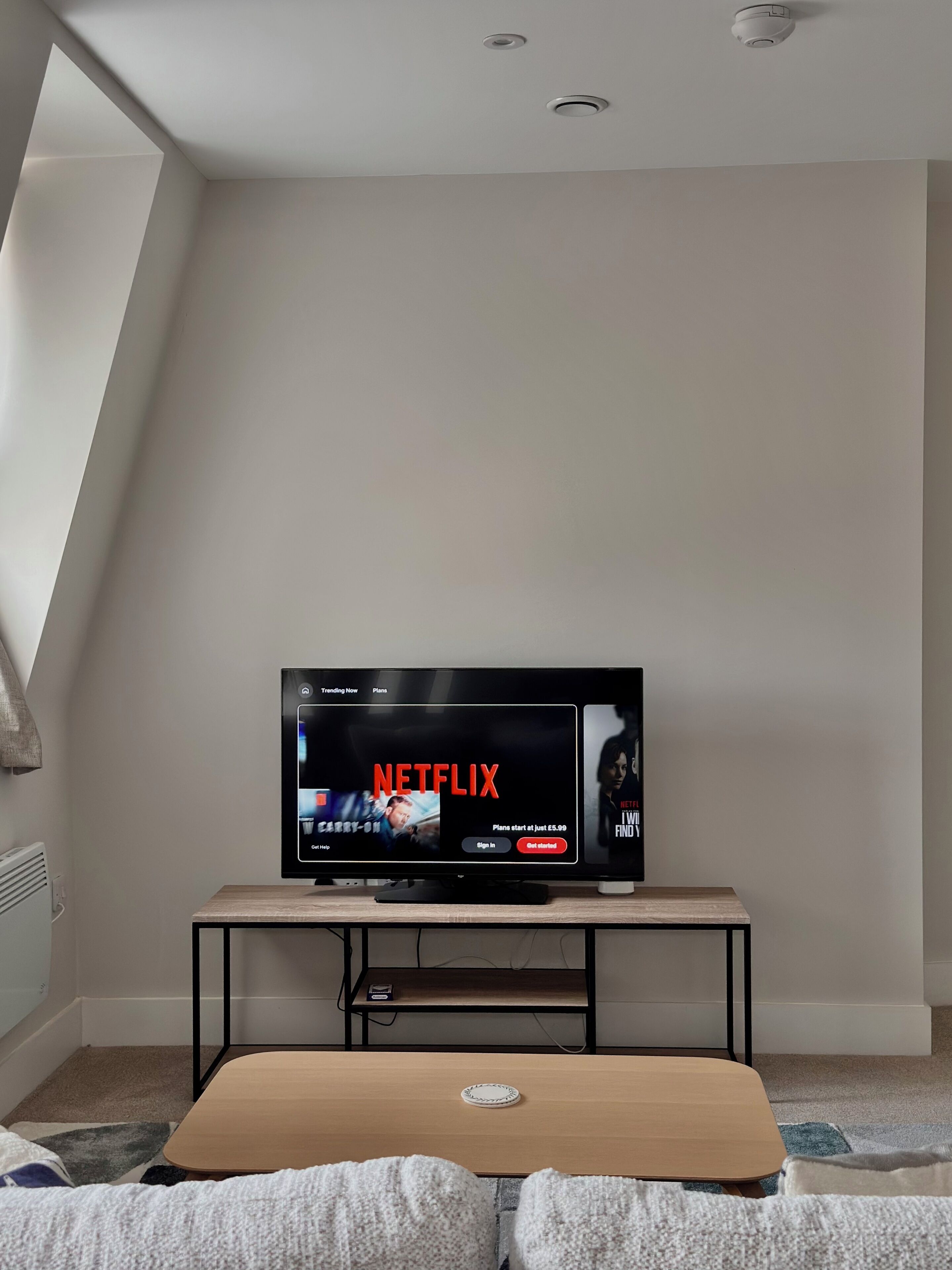 Un salon moderne avec un téléviseur à écran plat posé sur un meuble en bois, une petite table et un canapé recouvert d’un plaid.