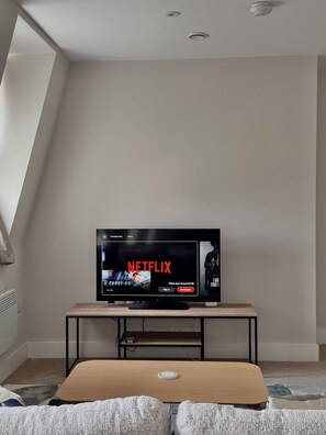 A modern living room with a flat-screen TV on a wooden stand, a small table, and a couch with a throw blanket.