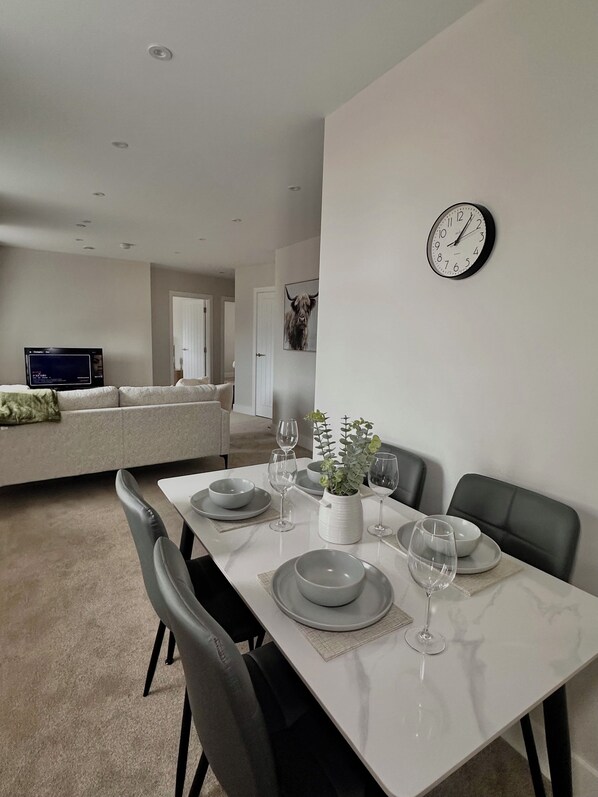 A dining area with a white table set for four, surrounded by black chairs. A clock is mounted on the wall above the table.