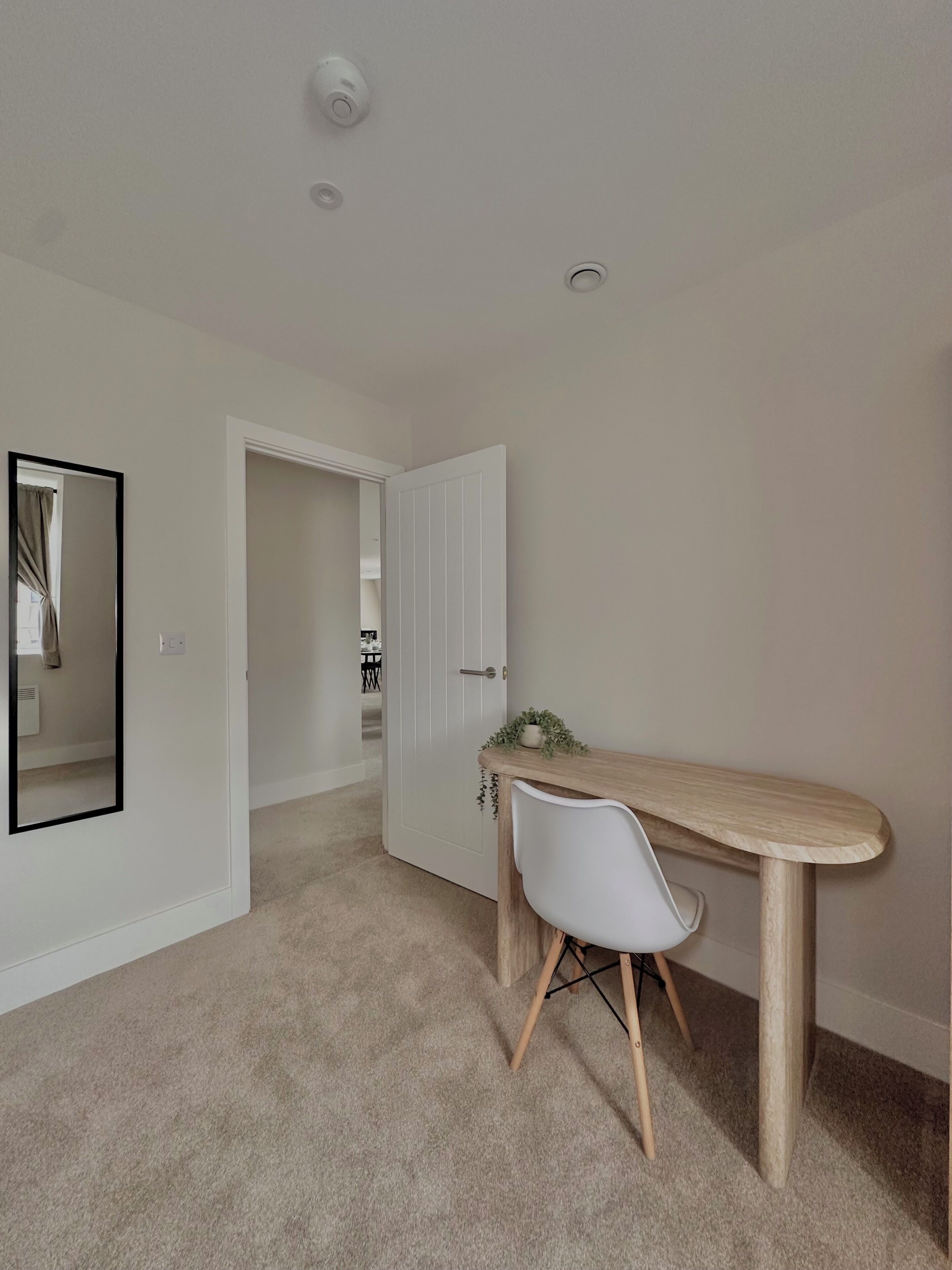 Une pièce avec un bureau en bois, une chaise blanche, un miroir et une porte donnant sur une autre pièce.