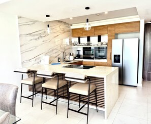 A modern kitchen with a marble backsplash, stainless steel appliances, and a central island with bar stools. - N/H Luxe 2BR/2.5 BA Villa Ocean Views Pool & Gym (Reading)