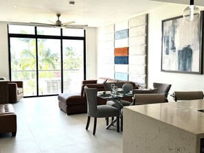 A modern living room with a sofa, dining table, and chairs, large windows with a view of palm trees, and a ceiling fan. - N/H Luxe 2BR/2.5 BA Villa Ocean Views Pool & Gym (Reading)