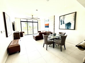 A spacious living area with a dining table set for four, a sofa, and a large window with a view of greenery. - N/H Luxe 2BR/2.5 BA Villa Ocean Views Pool & Gym (Reading)