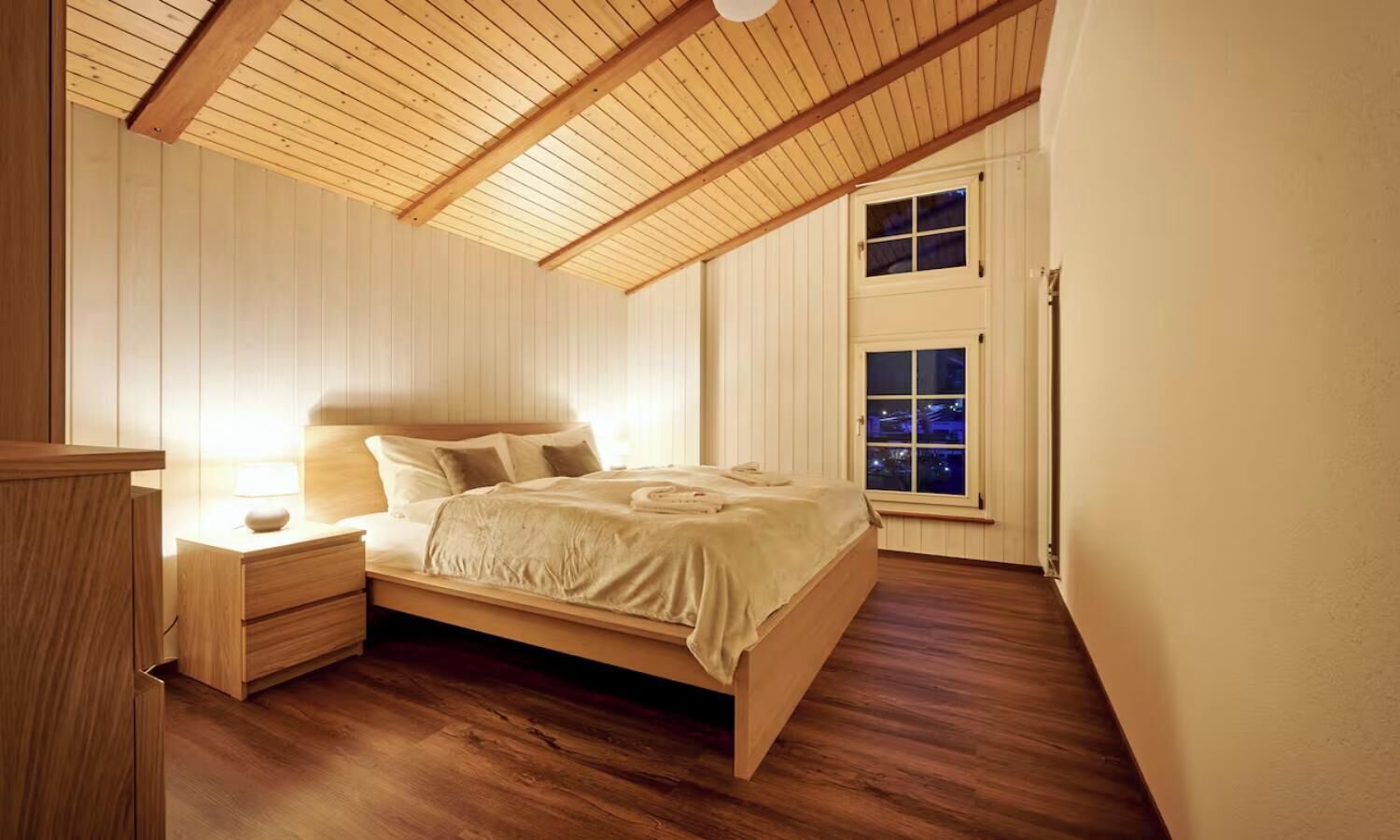 Une chambre avec un plafond en bois, un lit avec une tête de lit, une table de chevet et une fenêtre donnant sur la ville la nuit.