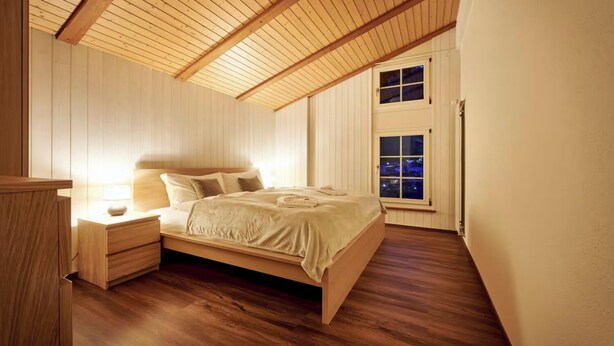 A bedroom with a wooden ceiling, a bed with a headboard, a nightstand, and a window with a view of the city at night. - CENTRAL 2-BEDROOM APARTMENT IN THE HEART OF LAUTERBRUNNEN (Lauterbrunnen)