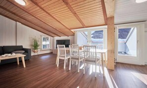 Ein geräumiges Zimmer mit Holzbalkendecke, einem Essbereich mit weißen Stühlen, einer Couch und einem großen Fenster mit Aussicht.