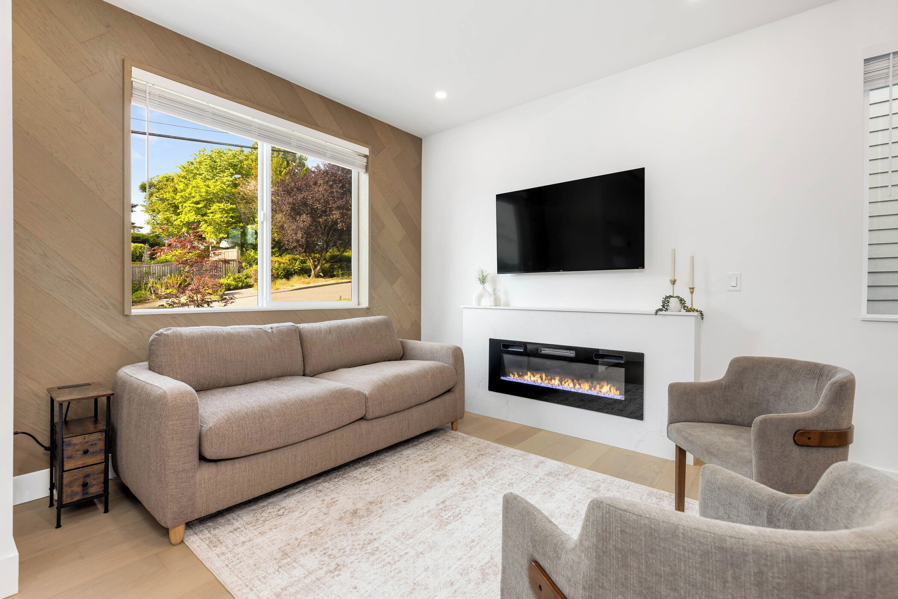 Ein modernes Wohnzimmer mit einem beigefarbenen Sofa, einem Kamin, einem Flachbildfernseher und Blick auf einen Garten durch ein großes Fenster.