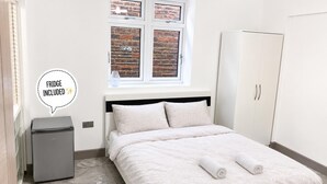 A bedroom with a bed, a wardrobe, a small fridge, and a window with a brick wall view. - Wembley Park - Free Parking (London)