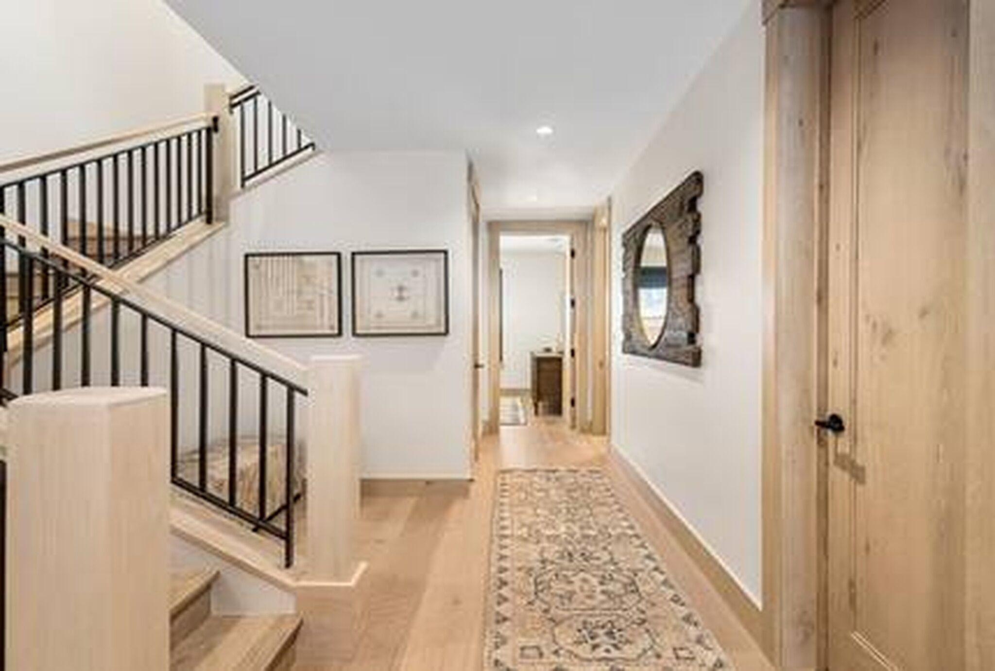 Un couloir doté d’un plancher en bois, d’un tapis, d’un escalier avec des rampes noires et d’une porte avec un miroir.