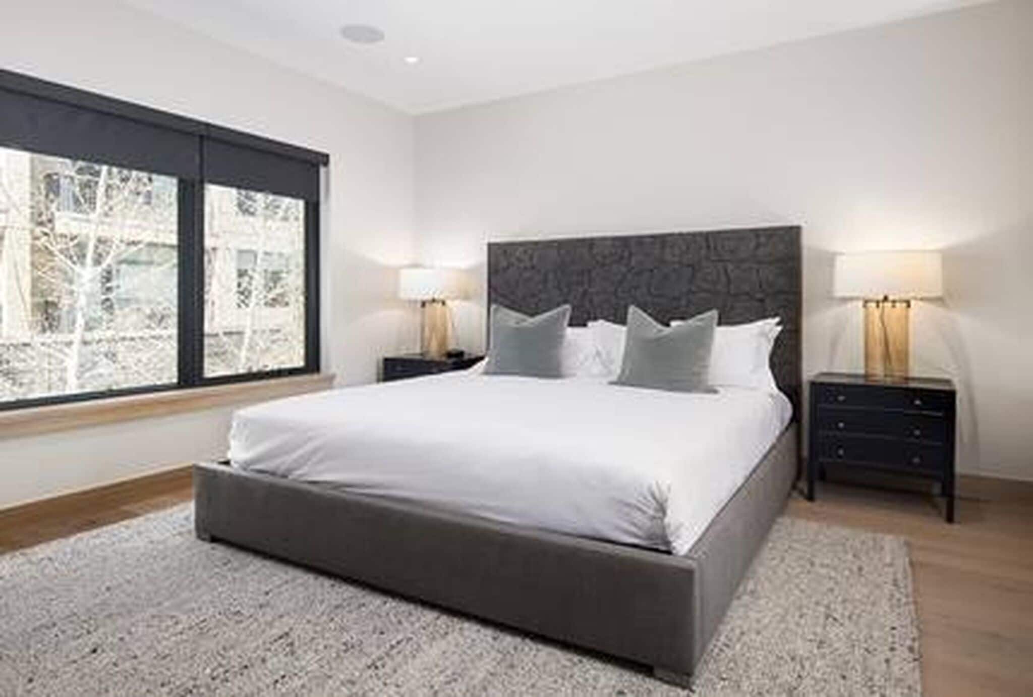 A modern bedroom with a large bed, two bedside tables, and a view through a window.