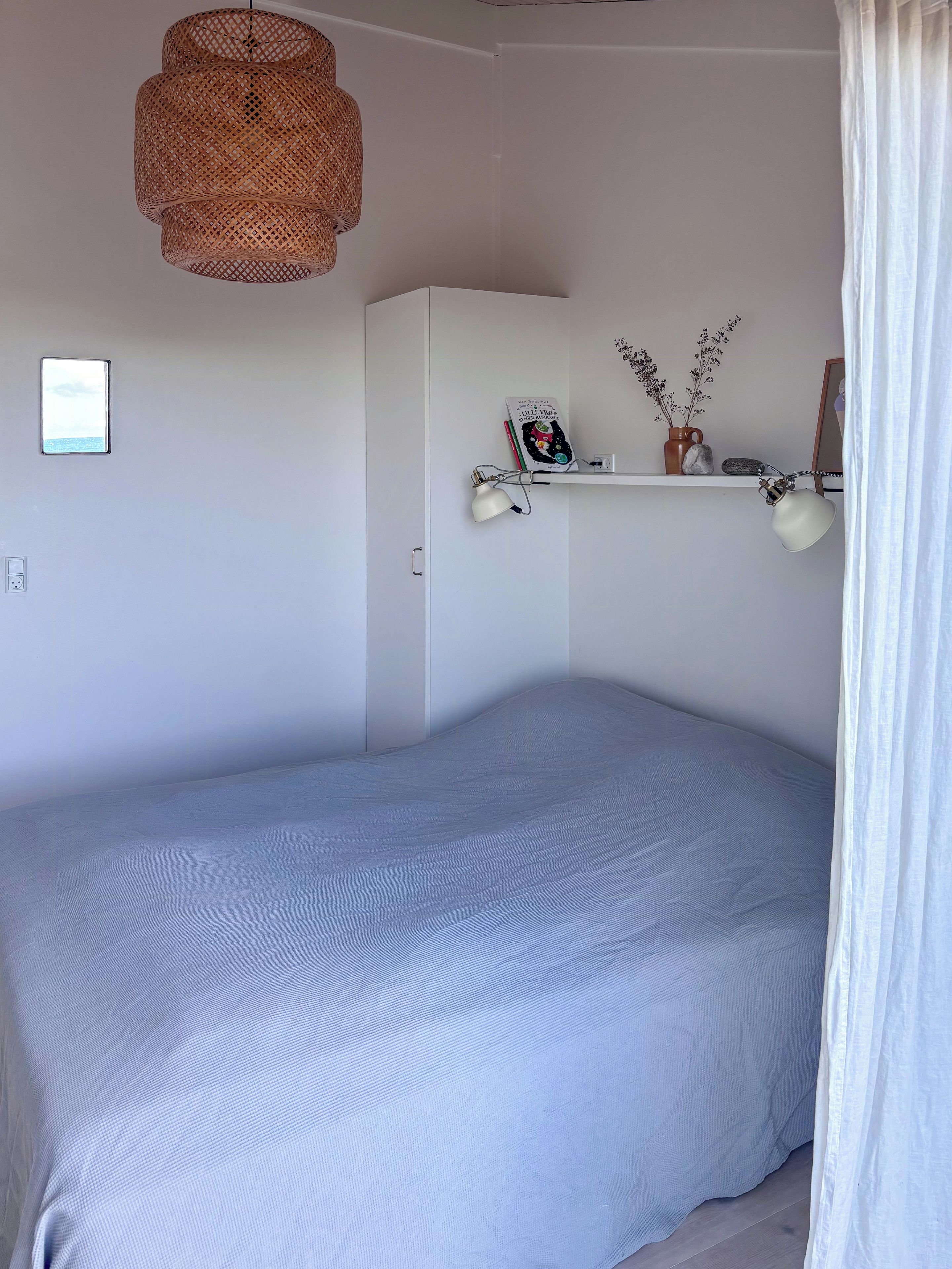 Ein Schlafzimmer mit einem Bett, einer weißen Wand, einem Regal mit Dekoration und einer gewobenen Leuchte.