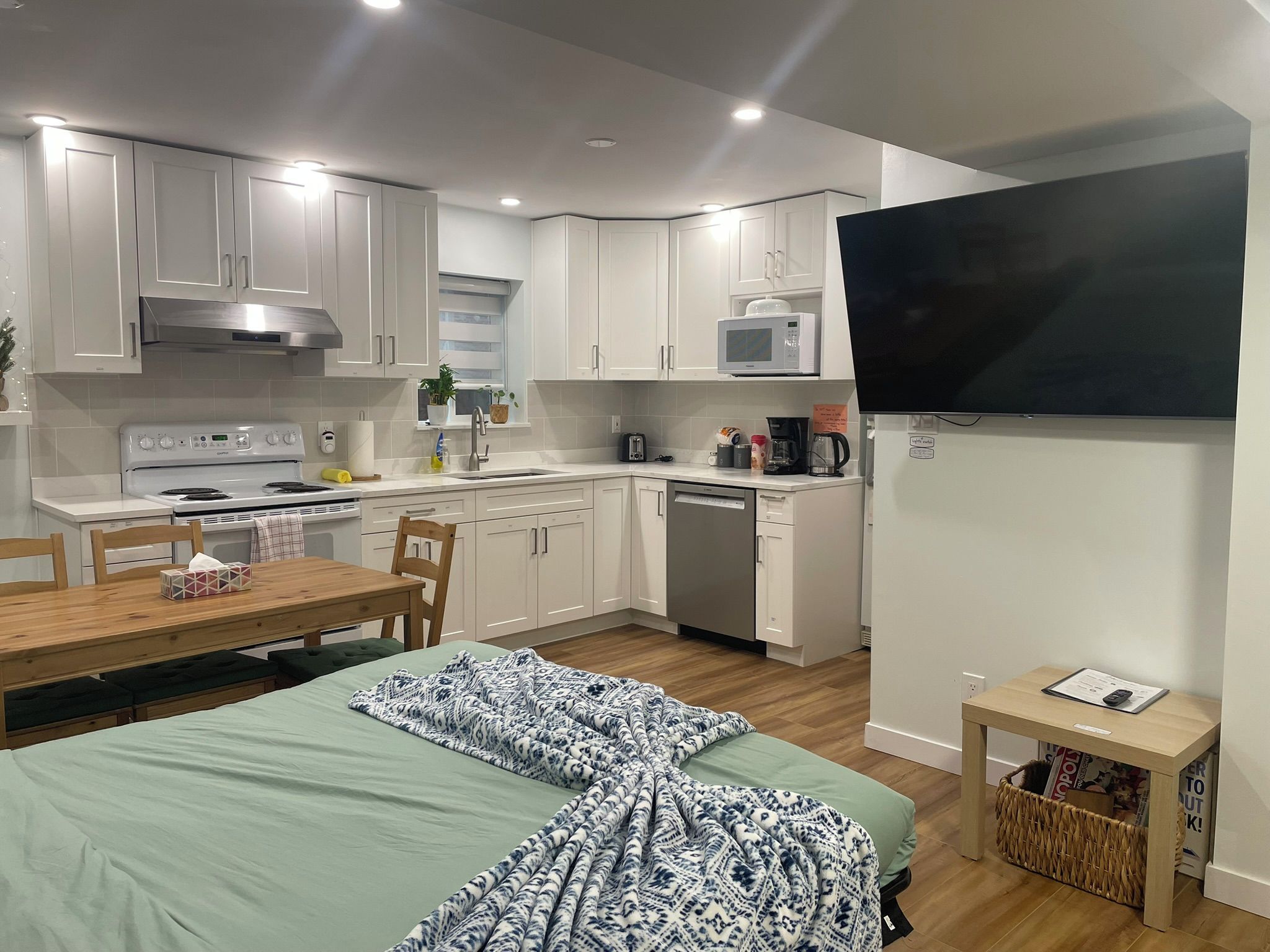 A compact kitchen with white cabinets, a microwave, a stove, and a dishwasher. There is a dining table with chairs and a bed with a patterned blanket.