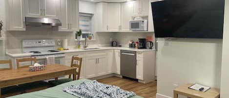 A compact kitchen with white cabinets, a microwave, a stove, and a dishwasher. There is a dining table with chairs and a bed with a patterned blanket.