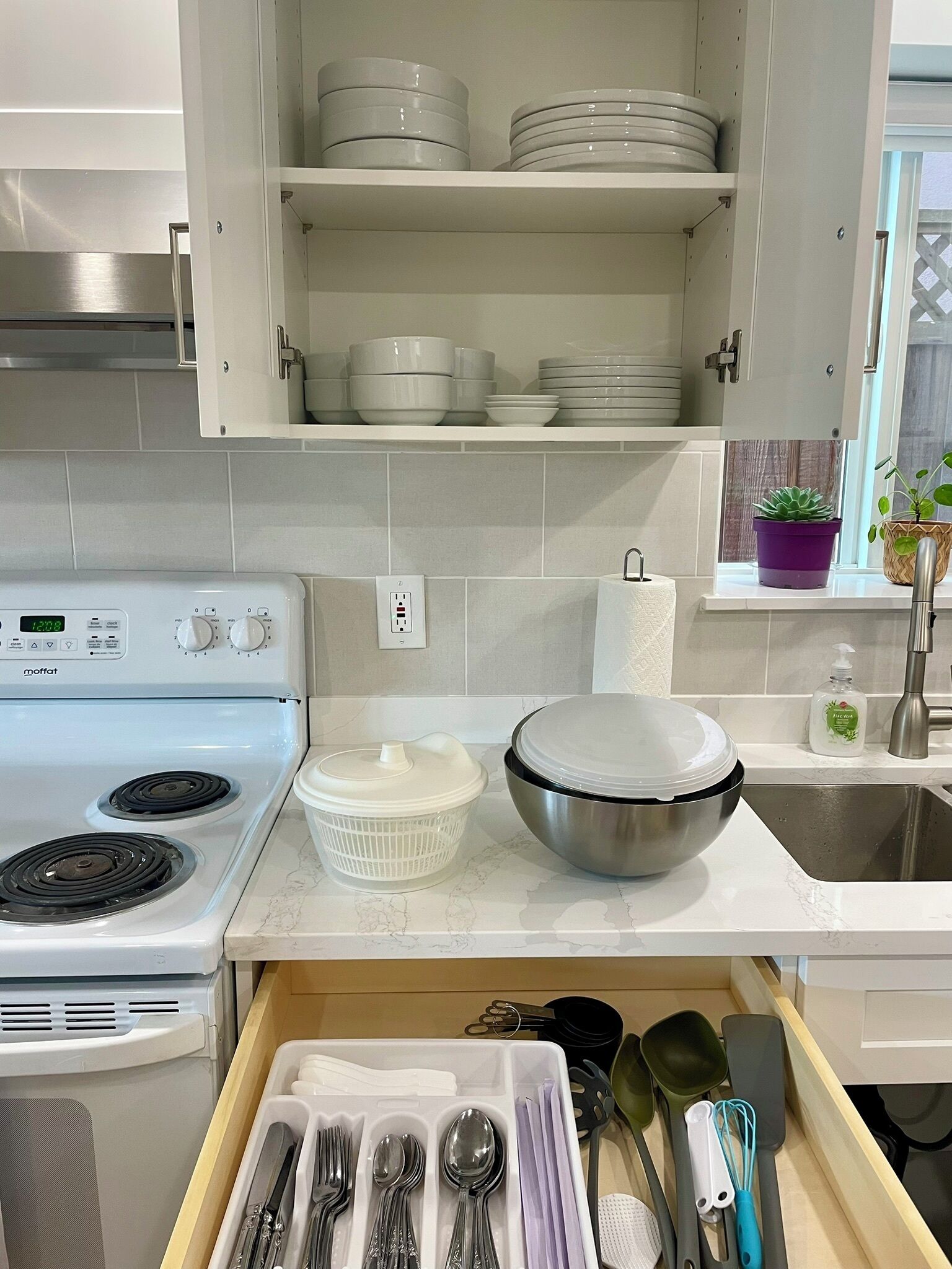 A kitchen with a white stove, a dish rack, a large mixing bowl, and a drawer with utensils.