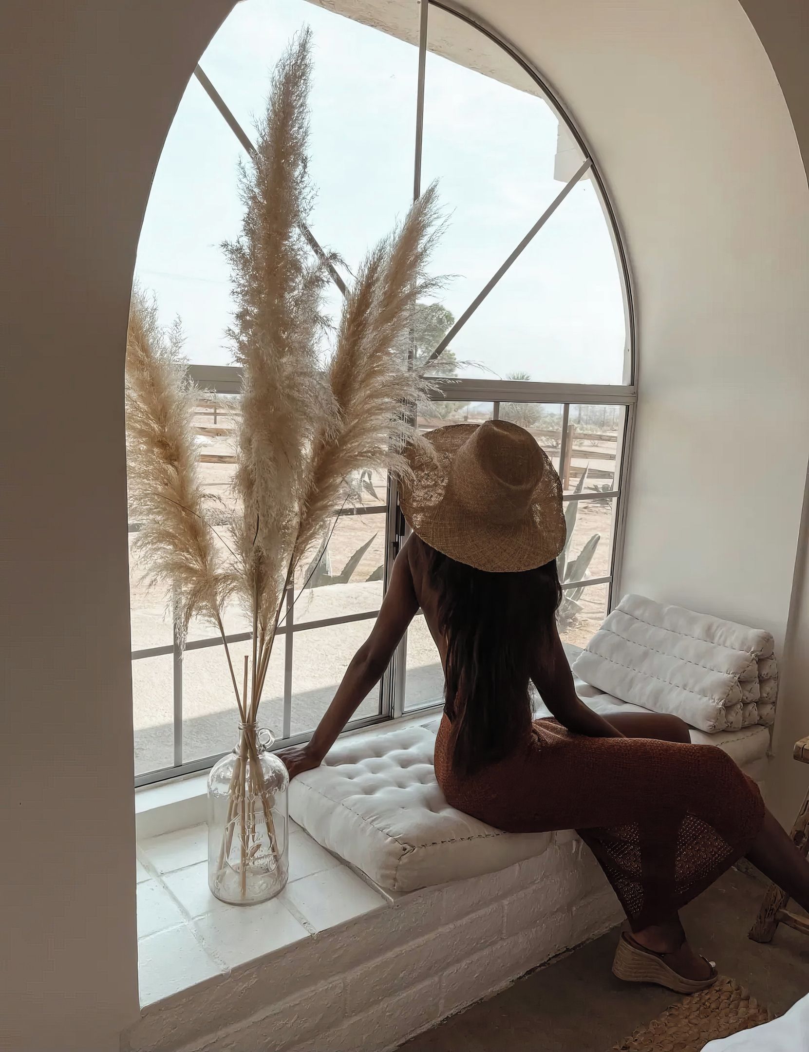 A person sitting by a large arched window with pampas grass, a white cushioned bench, and a clear glass vase with reeds.