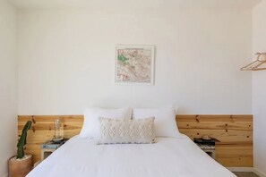 Ein Schlafzimmer mit einem hölzernen Kopfende, einem Bett mit weißer Bettwäsche, einer Karte an der Wand, einer Kakteenpflanze und einem Nachttisch mit Lampe und Buch.