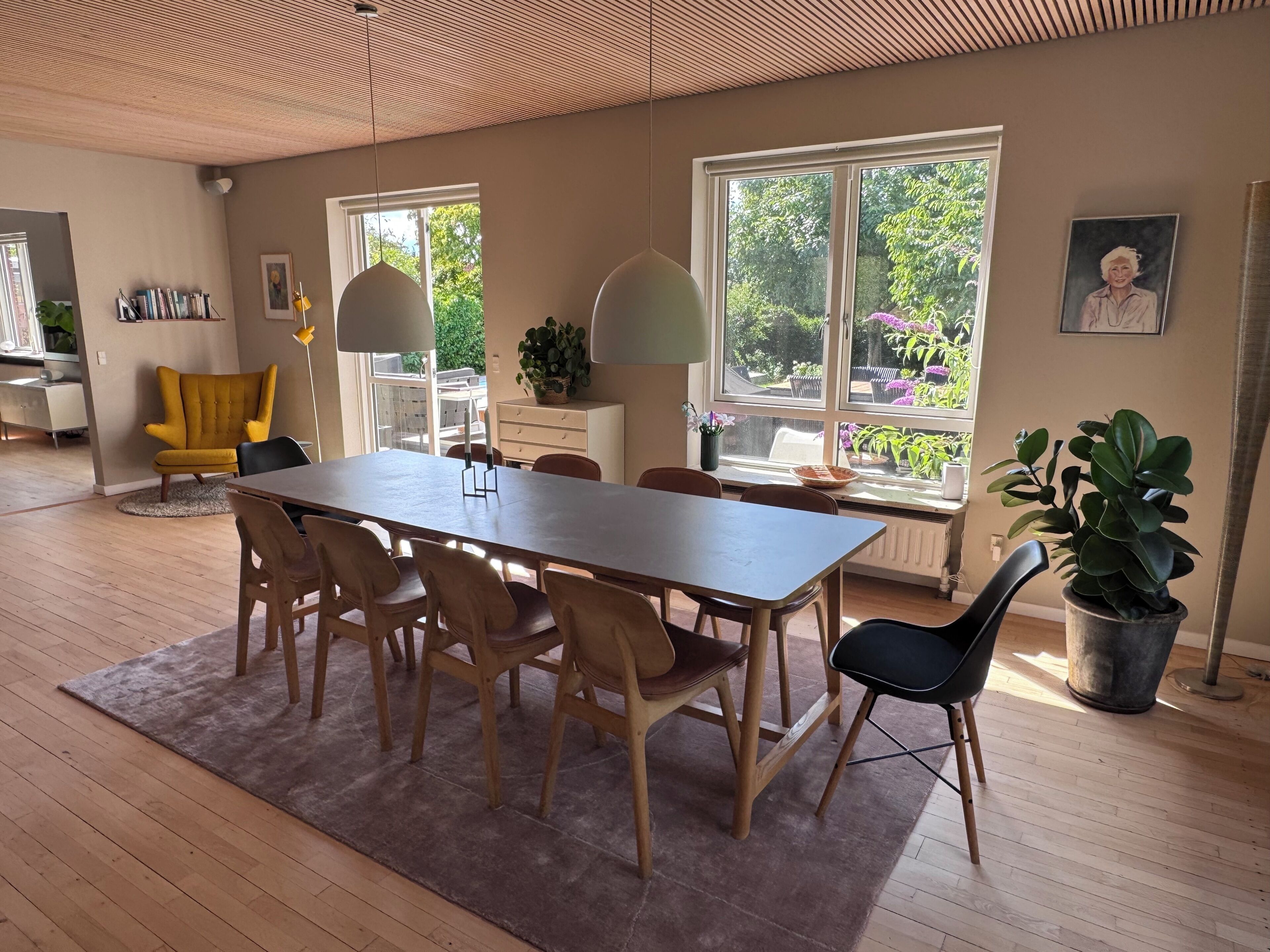 Un espace repas avec une longue table, des chaises et un tapis. De grandes fenêtres offrent une vue sur la verdure environnante.