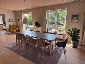 Ruokailutilassa on pitkä pöytä, tuoleja ja matto. Ikkunoista avautuu näkymä vehreään maisemaan.