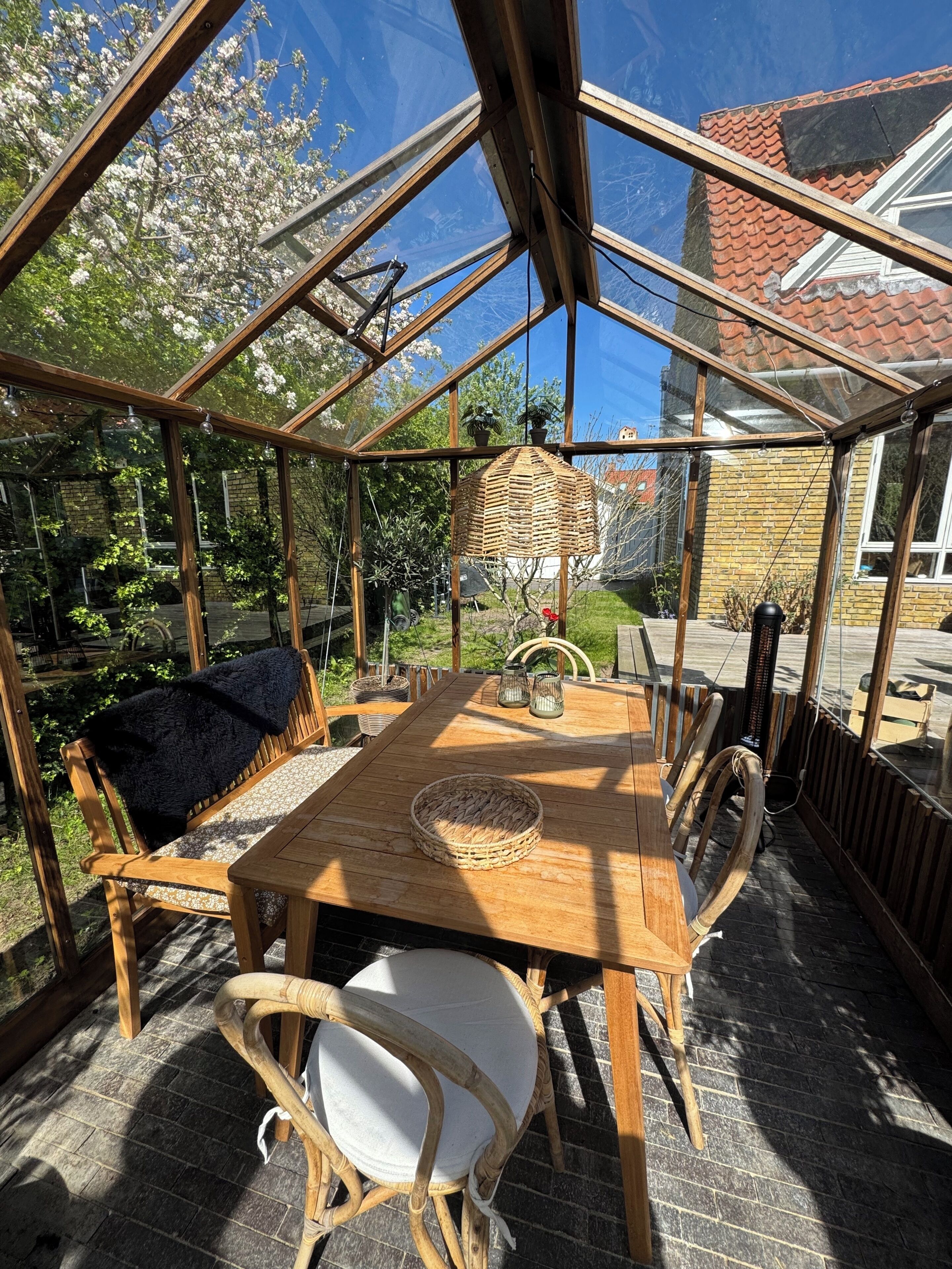 Un espace extérieur abrité, avec une table et des chaises en bois, une lampe en osier suspendue, et une vue sur un jardin et une maison en arrière-plan.