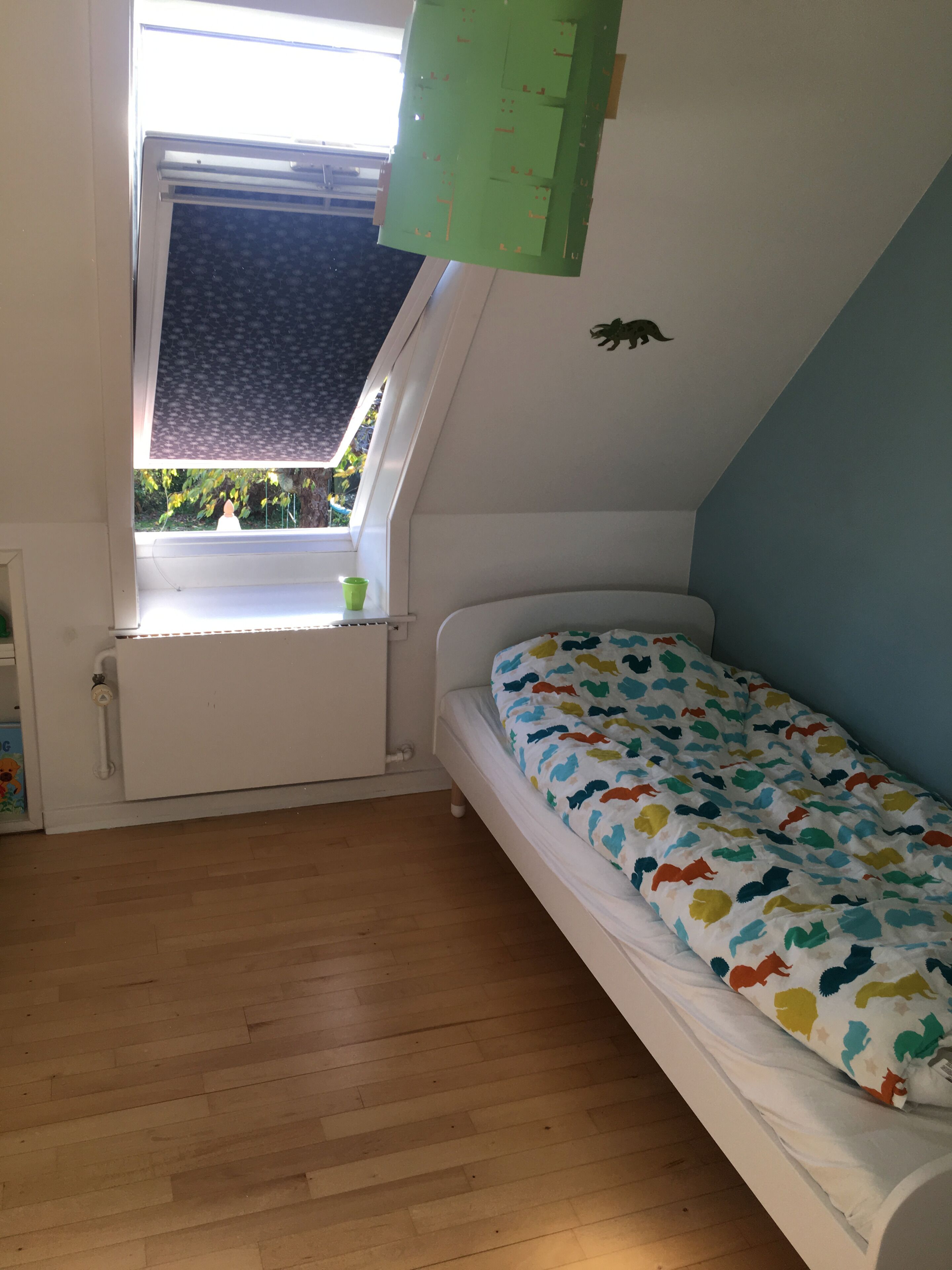 Une pièce avec un plafond en pente, un lit recouvert d’une courtepointe à motifs animaliers colorés, une fenêtre donnant sur un paysage verdoyant et une armoire verte.