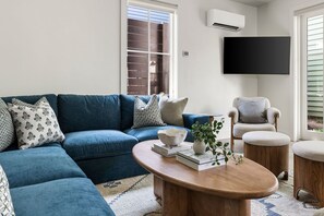 Una sala de estar con un sofá azul, una mesa de centro redonda, un sillón gris y un televisor de pantalla plana montado en la pared.