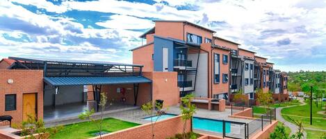 A modern multi-story residential building with a swimming pool, covered parking, and a landscaped garden.