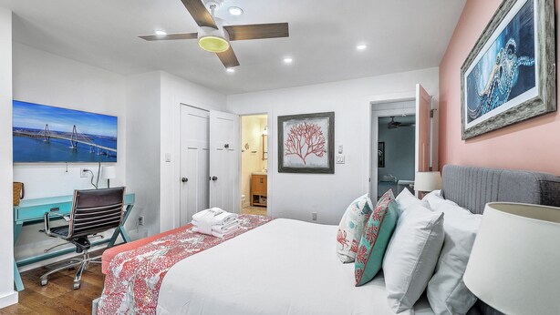 Quarto de hotel com cama, escrivaninha, cadeira, ventilador de teto e obras de arte nas paredes.