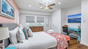Quarto com cama, escrivaninha, cadeira, ventilador de teto e um quadro na parede.