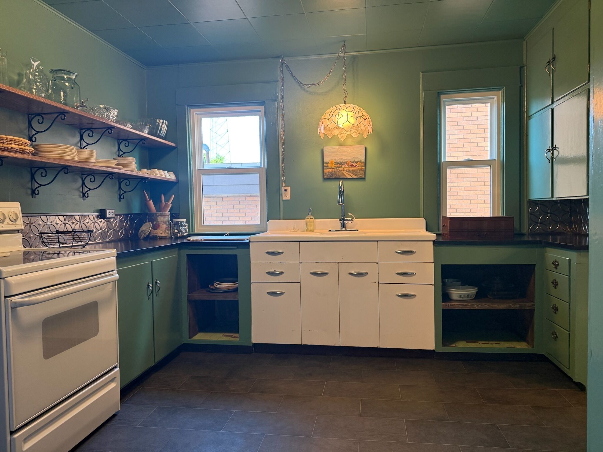 Cocina con estufa, horno y alacenas. Hay dos ventanas y una lámpara colgante.