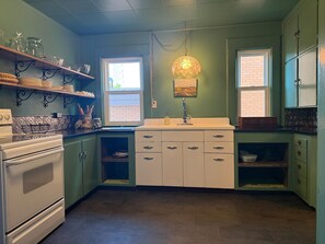 Une cuisine équipée d’une cuisinière, d’un four et de placards. Elle comporte deux fenêtres et un luminaire suspendu.