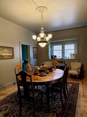 A dining area with a wooden table, chairs, and a chandelier. There is a window with a view of the outside.