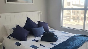 A bedroom with a bed, pillows, a blanket, a painting on the wall, and a window with a view. - Modern 2-bedroom apartment in Langebaan near the beach (Langebaan)
