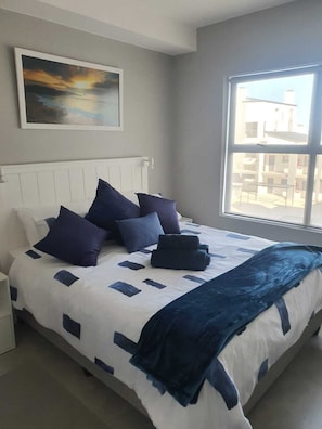 Ein Schlafzimmer mit einem Bett, Kissen, einer Decke, einem Bild an der Wand und einem Fenster mit Blick.