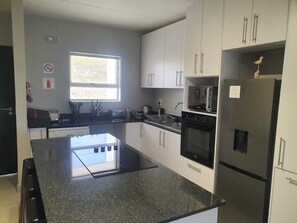Una cocina moderna con armarios blancos, encimera negra y electrodomésticos de acero inoxidable, incluyendo un refrigerador, horno y microondas.