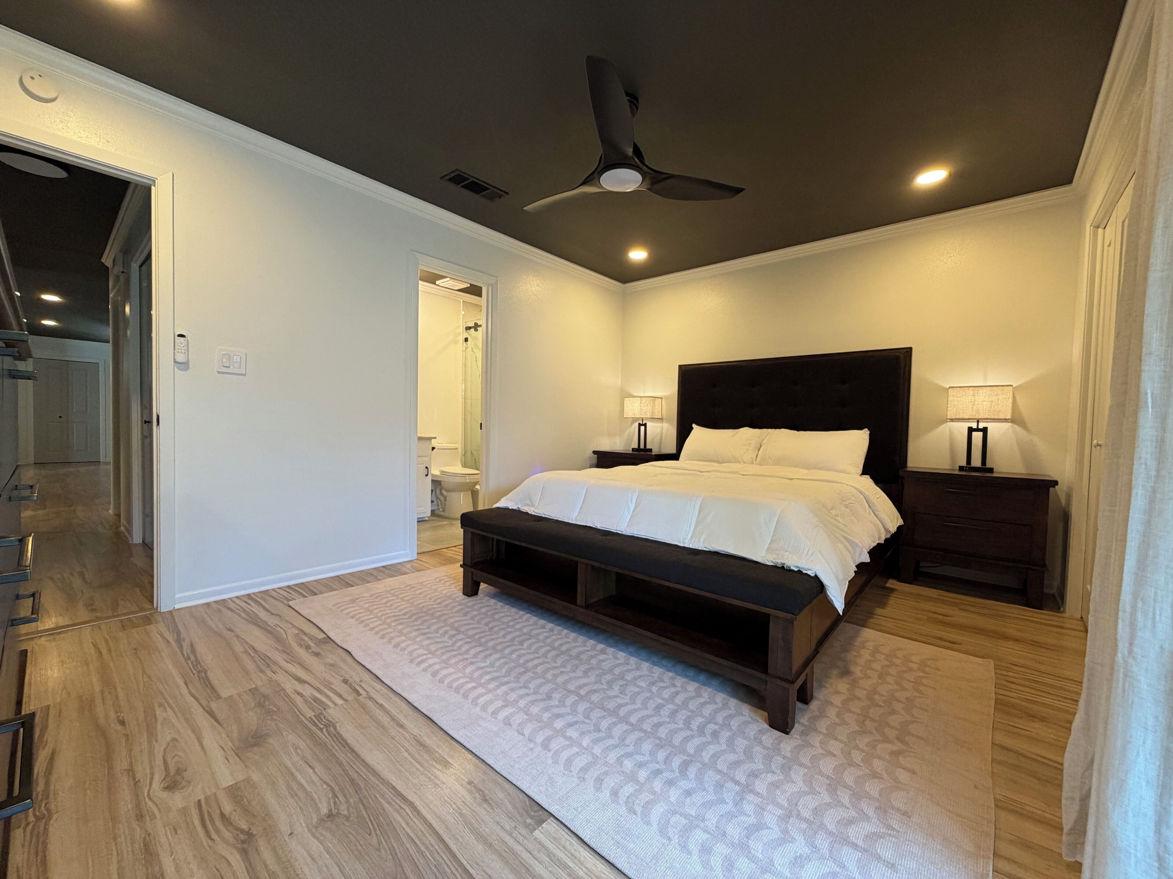 Un dormitorio con cama, mesitas de noche, ventilador de techo y un baño visible a través de una puerta abierta.