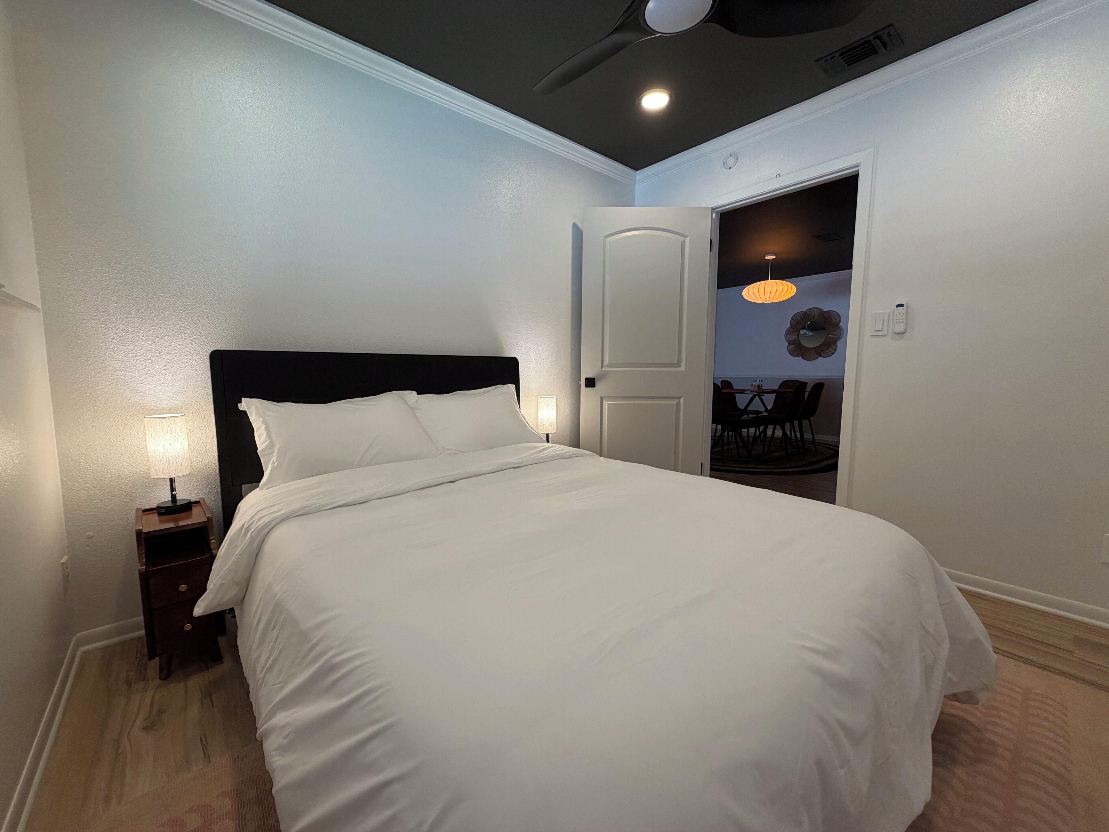 A neatly made bed with white linens in a room with a dark ceiling and a door leading to a dining area.