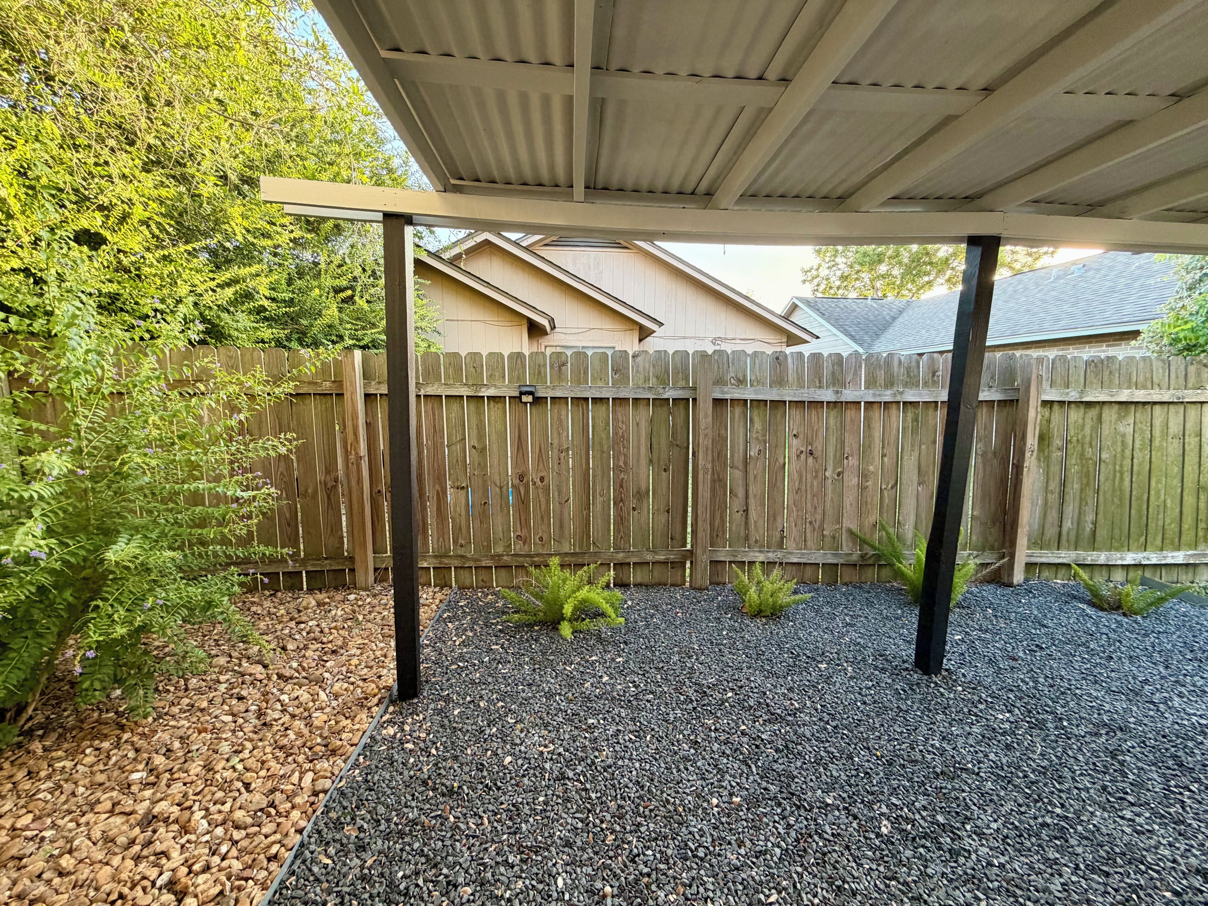 A covered outdoor area with a wooden fence, gravel ground, and a metal roof structure.