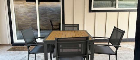 Un patio exterior con una mesa de madera y cuatro sillas, una puerta corrediza de vidrio y una ventana con persianas.
