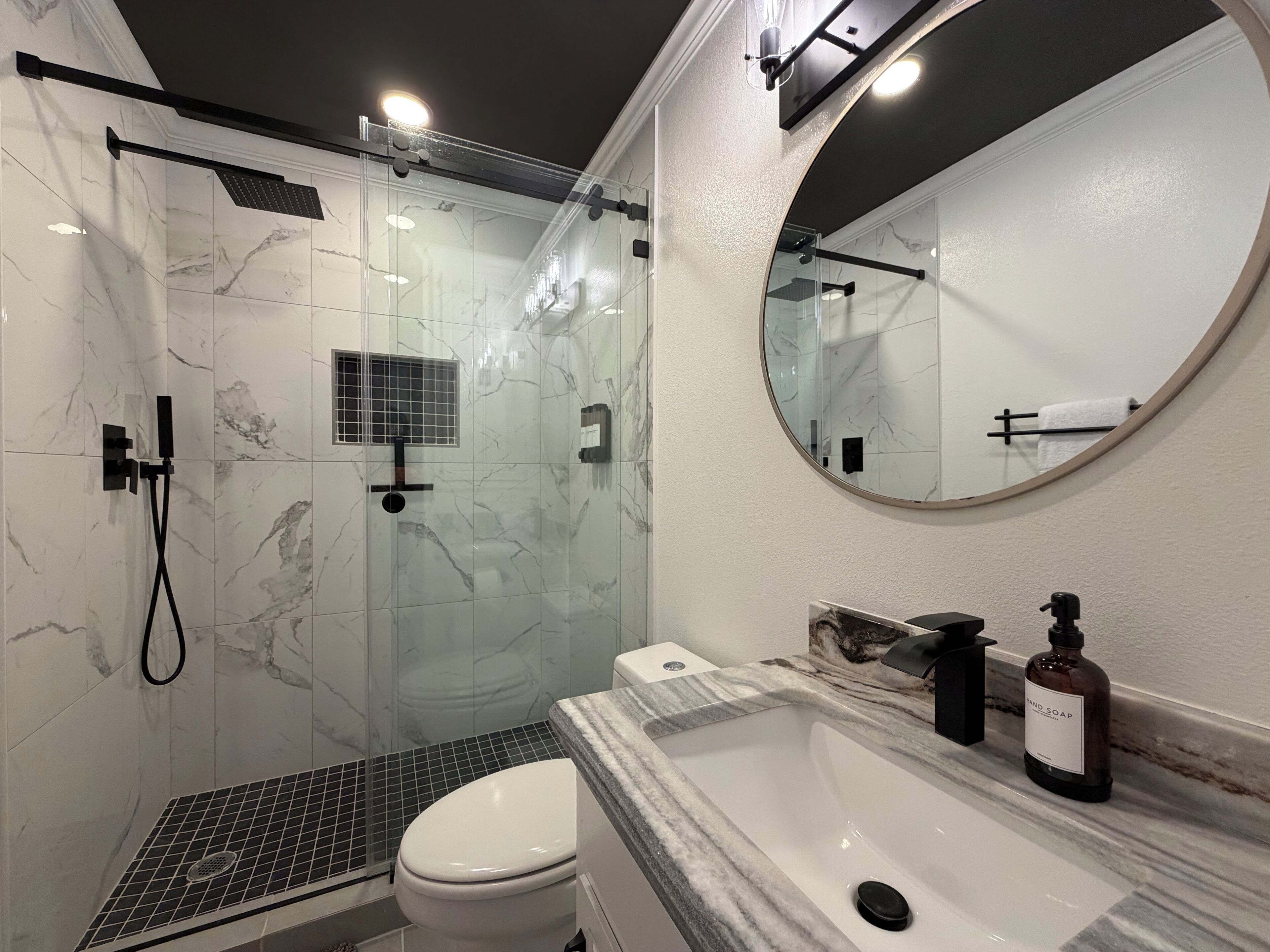 Ein modernes Badezimmer mit einer gläsernen Dusche, einer weißen Toilette, einem мраморном Waschbecken und einem runden Spiegel.