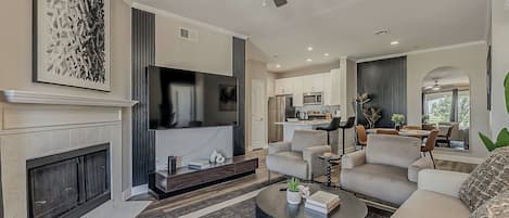 A modern living room with a fireplace, a flat-screen TV, a ceiling fan, and a dining area with a kitchen in the background.