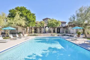 Piscina com espreguiçadeiras e guarda-sóis, cercada por árvores e um edifício com entrada coberta.